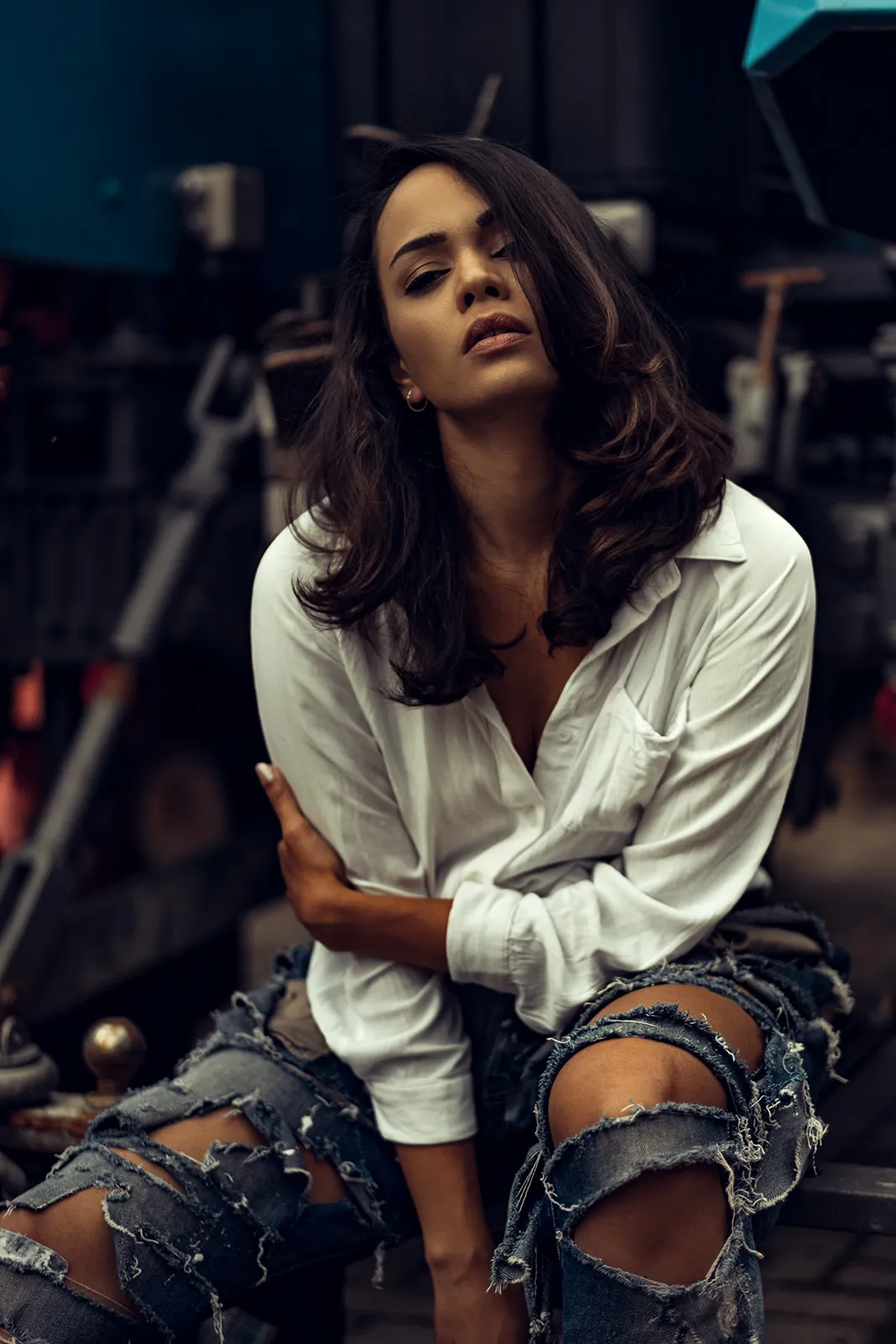 Dark-haired young woman in a white linen shirt sits with crossed arms against an industrial backdrop.
