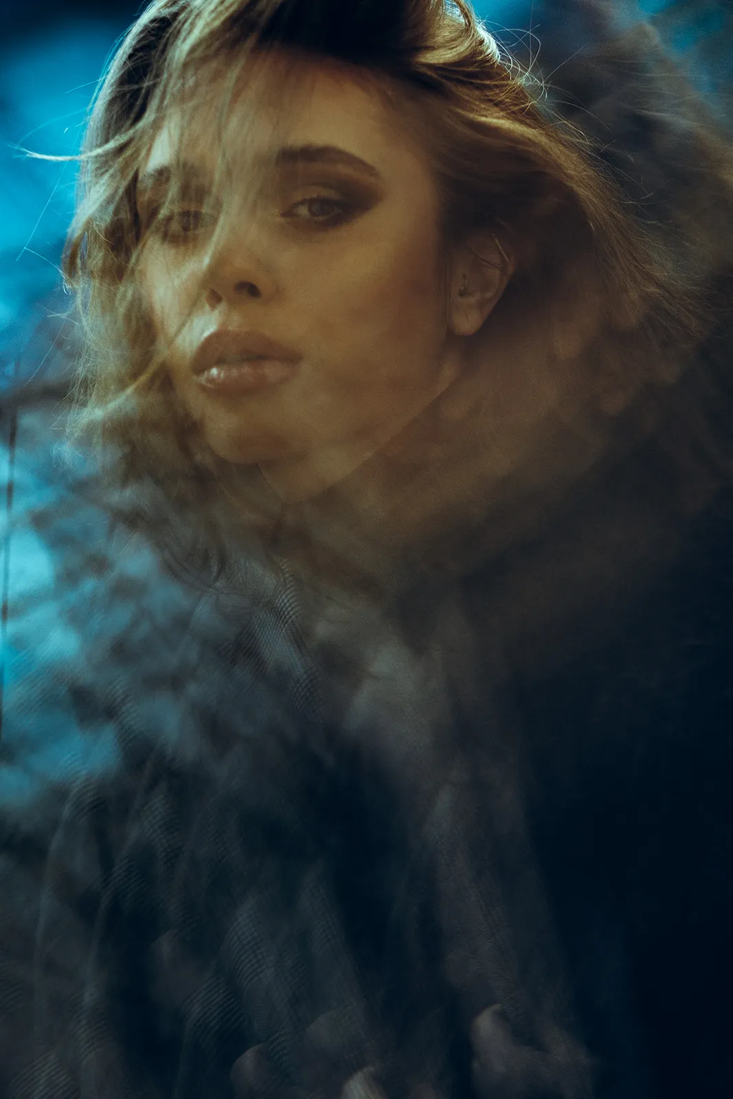 A young woman's face emerges from darkness through wind-swept hair and cool blue backlight.
