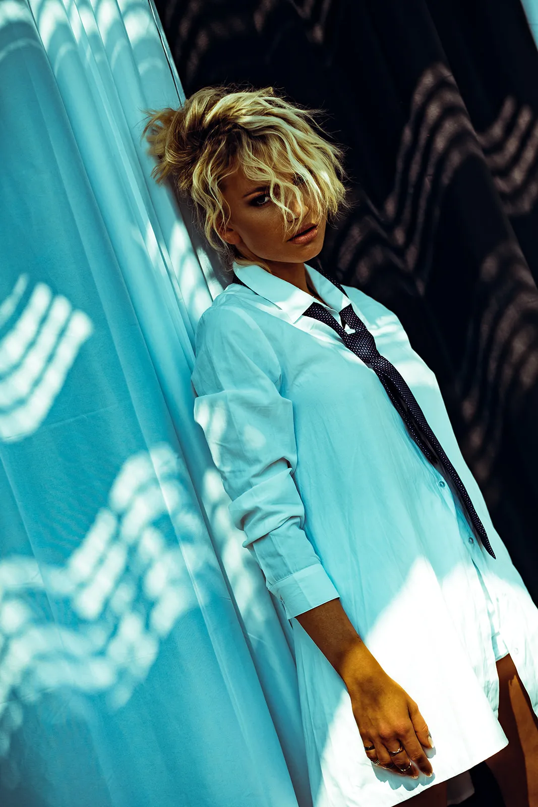 Blonde woman in a light blue oversized shirt leaning against a curtain in backlight.