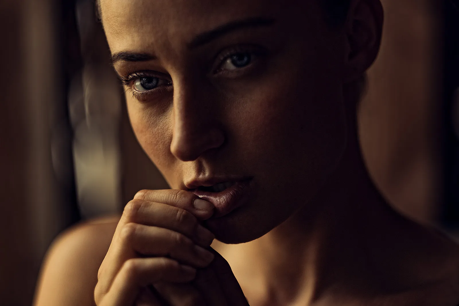 Close-up of a young woman in warm shadow light thoughtfully holding her hands to her lips.