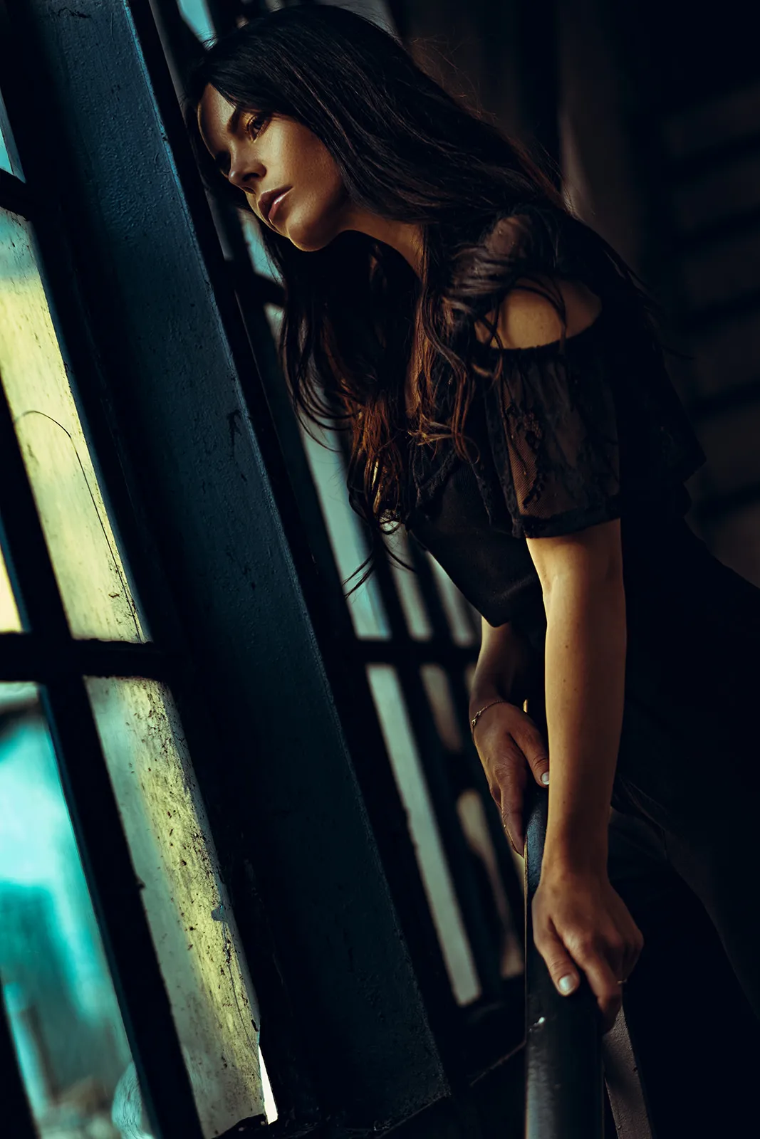 Young woman leaning over a railing gazing through barred industrial windows into green-teal light.