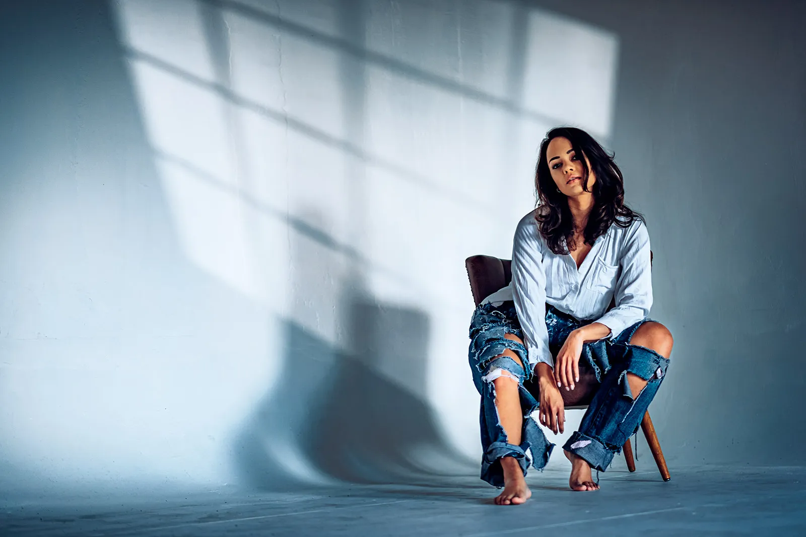 Barefoot on a chair, she gazes upward in ripped jeans and a loose white shirt.