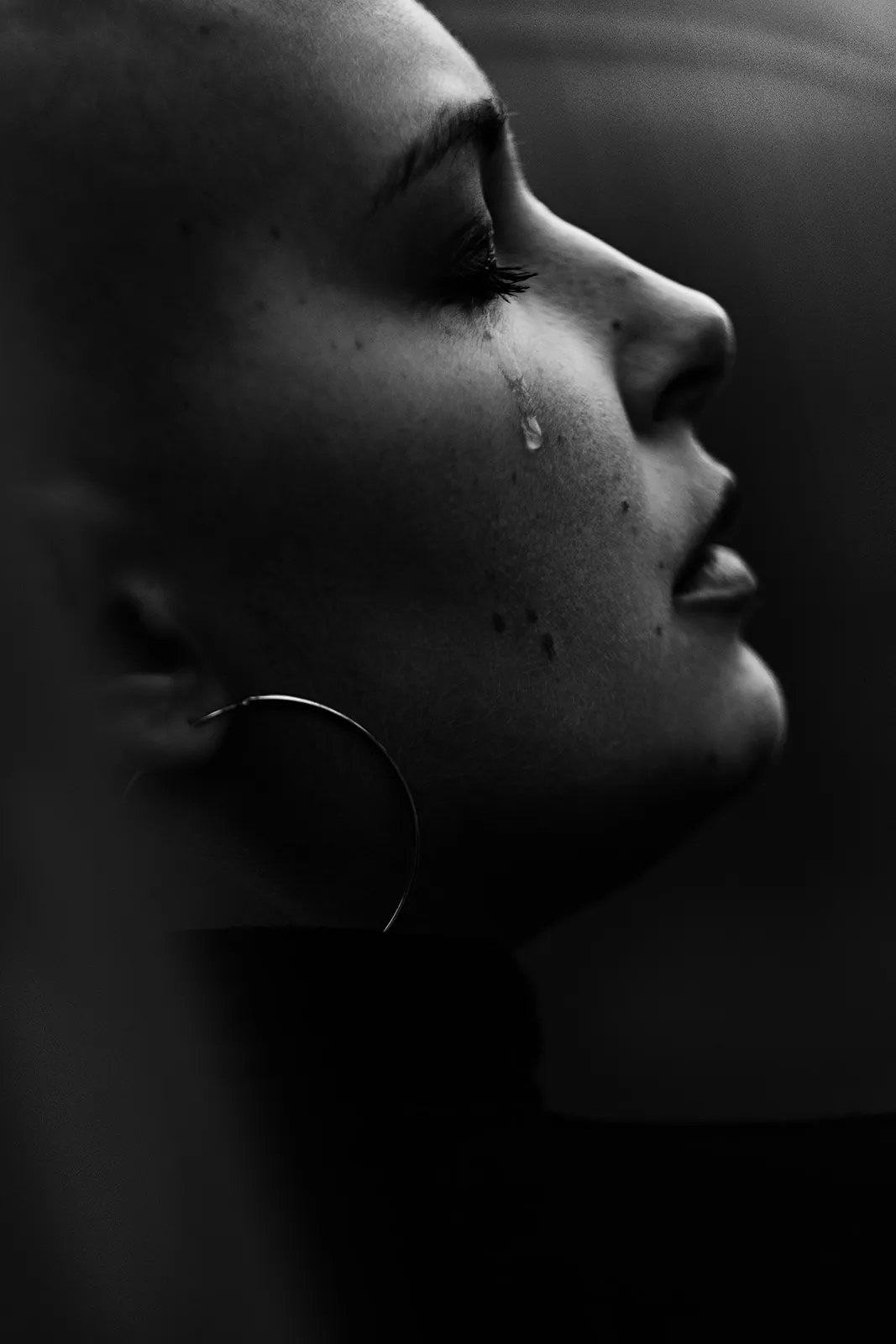 Side profile with closed eyes and a single tear on the cheek against dark backlight.