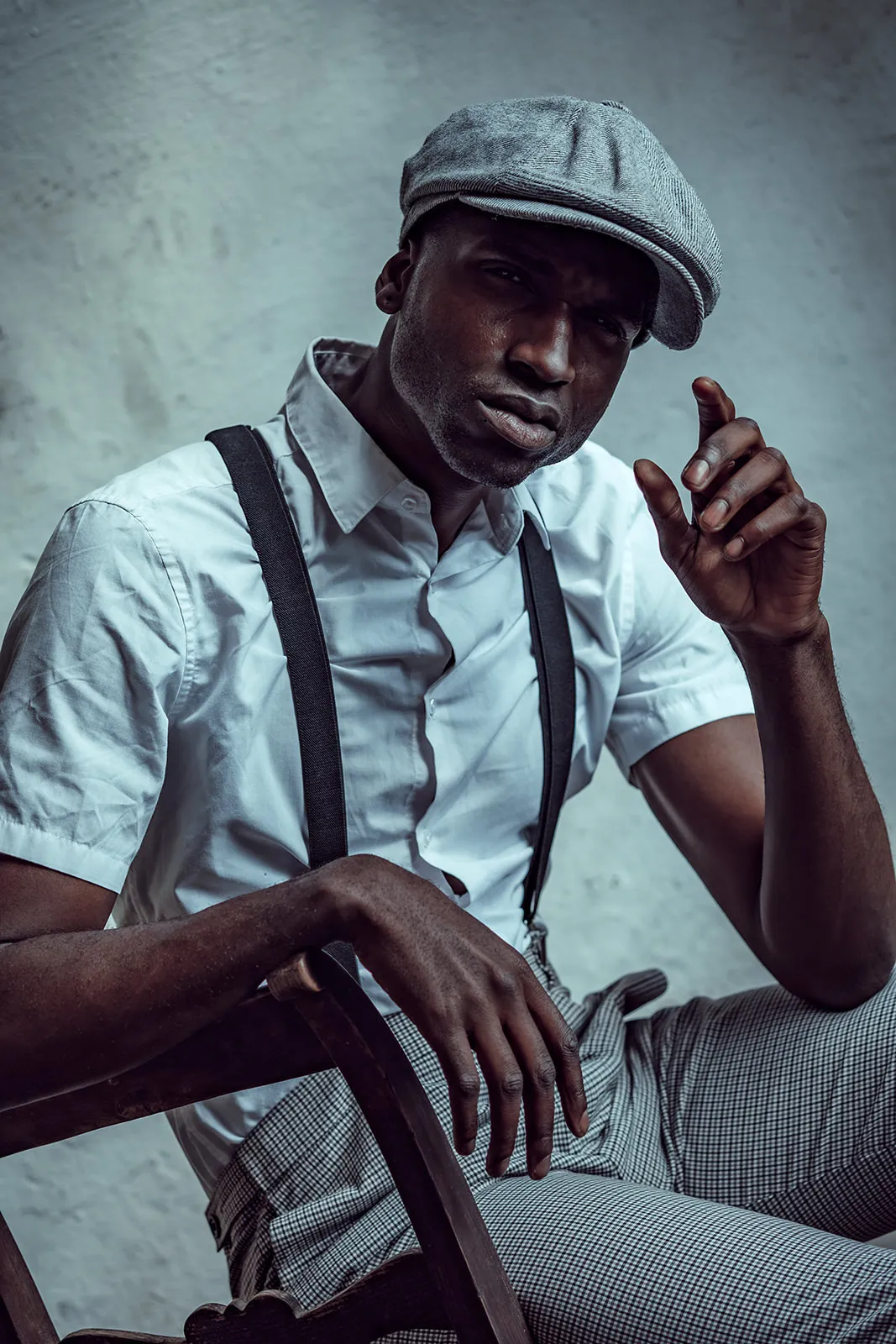 A man in a white short-sleeve shirt and suspenders sits quietly on a wooden chair holding a steady gaze toward the camera.