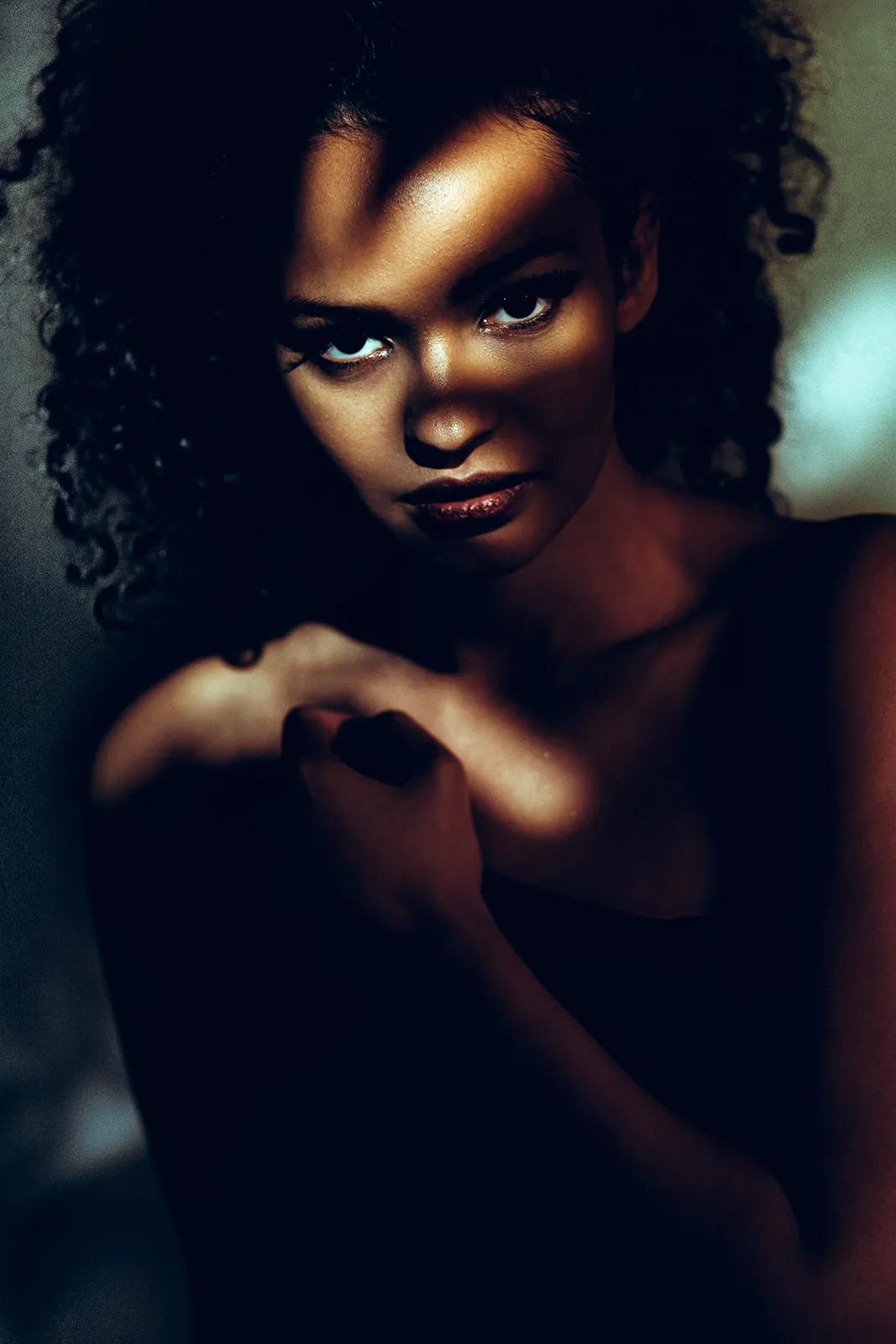 Warm raking light falls across a face with an intense gaze and crossed hands against a dark background.