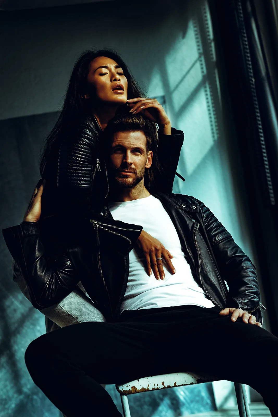A woman in a black leather jacket leans over a seated man, resting her hand on the back of his neck.