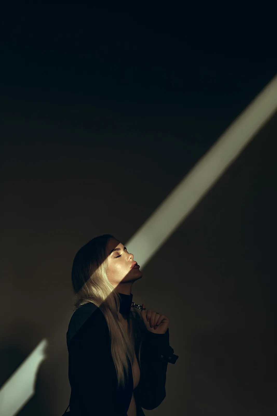 A narrow beam of light falls diagonally across an upturned face with closed eyes in near darkness.