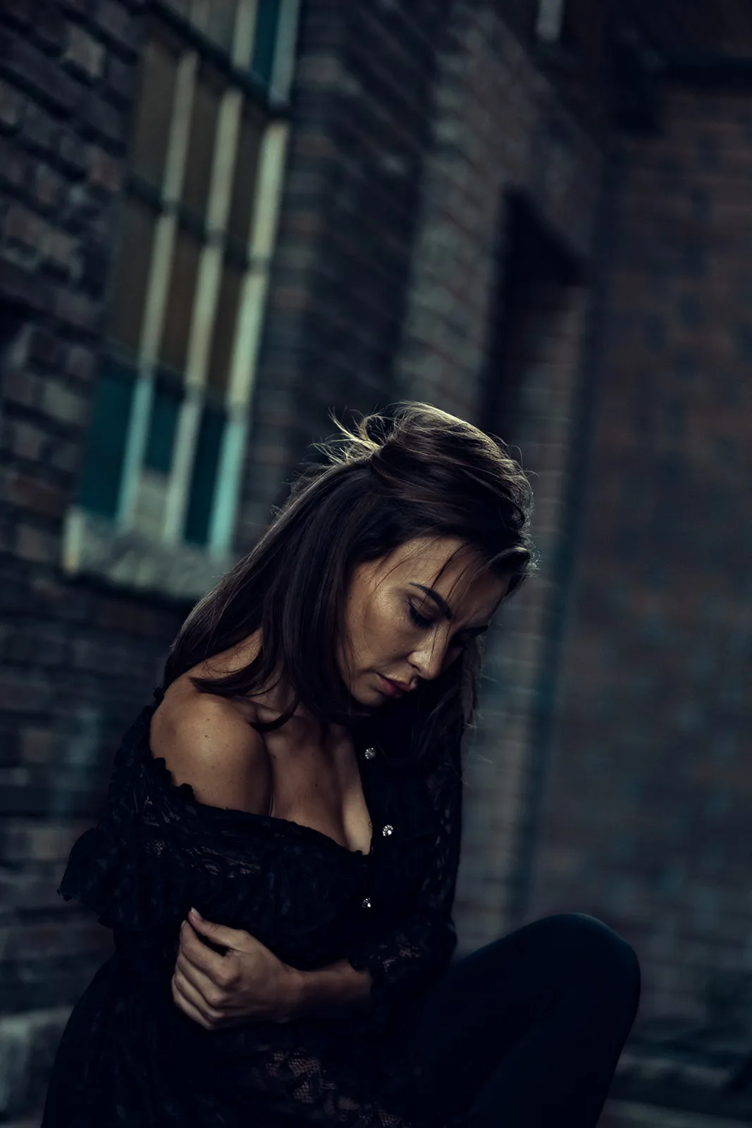 Young woman in a black lace top seated against a brick wall, gaze cast downward.