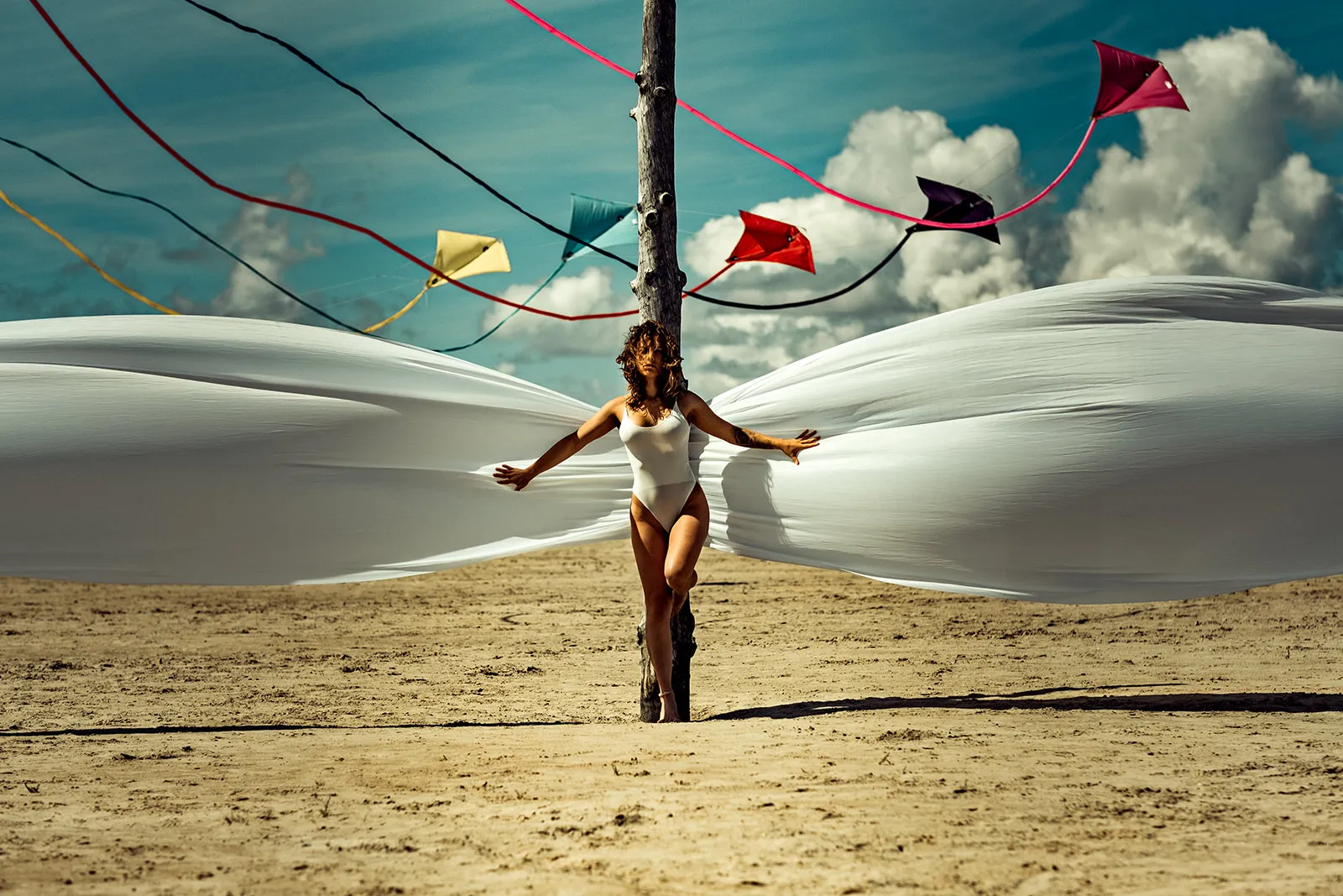 A woman in a white swimsuit stands at a wooden pole as white fabric billows outward on both sides in the wind.